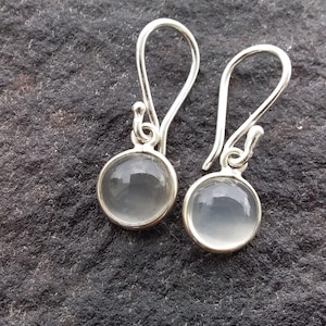 May include: A pair of silver earrings with a round, clear, gray stone set in a silver bezel. The earrings have a simple hook design. The background is a dark, textured surface.