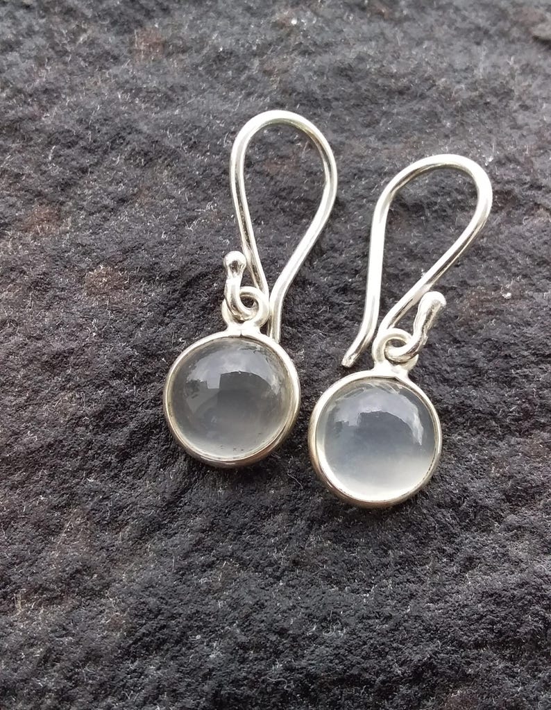 May include: A pair of silver earrings with a round, clear, gray stone set in a silver bezel. The earrings have a simple hook design. The background is a dark, textured surface.