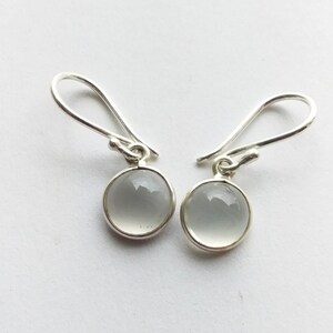 May include: A pair of silver drop earrings with round, pale gray, polished stones. The stones are set in a silver bezel and hang from a simple silver hook. The earrings are displayed against a white background.
