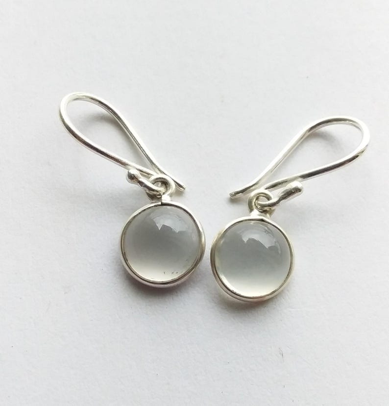 May include: A pair of silver drop earrings with round, pale gray, polished stones. The stones are set in a silver bezel and hang from a simple silver hook. The earrings are displayed against a white background.