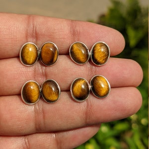 Pode incluir: Um par de brincos de prata com pedras de olho de tigre de forma oval. Os brincos são mostrados em uma mão.