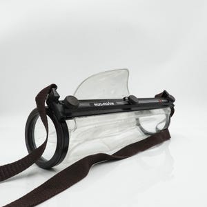 May include: A clear, waterproof camera housing with a black frame and a brown shoulder strap. The housing has a clear, curved lens and a black ring around the lens. The text "ewa-marine" is printed on the top of the housing.