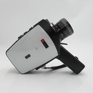 May include: A vintage black and silver film camera with a prominent lens and a handle. The camera has a silver body with black accents and a black lens. The lens has measurement markings. A black strap is attached to the camera.