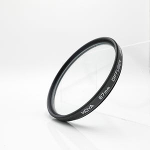 May include: A black Hoya 67mm diffuser filter with the word "DIFFUSER" printed on the side. The filter is made of clear glass and has a black metal ring. The filter is angled slightly, with a white background.