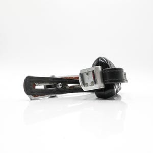 May include: A vintage black leather camera wrist strap with a metal and black camera attachment. The strap is attached to a metal piece with a small dial and a black plastic camera body.
