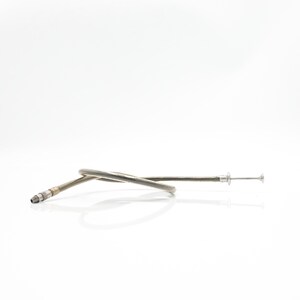 May include: A silver-colored, flexible cable with a threaded end and a small, button-like component at the opposite end. The cable is curved and lies on a white surface.