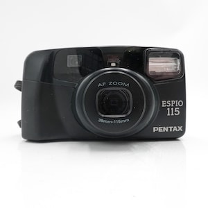 以下が含まれることがあります： AFズームと38mm-115mmレンズを備えた黒色のPentax Espio 115フィルムカメラ。カメラには内蔵フラッシュがあり、「Espio 115」と「Pentax」の文字が確認できます。