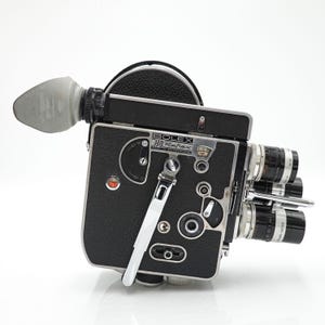 Op de afbeelding: Een vintage Bolex H8 Reflex filmcamera. De camera is zwart met zilveren accenten en meerdere lenzen. Hij heeft een zoeker, wijzerplaten en een handvat. De camera is ontworpen voor 8 mm film, een klassieker in de filmwereld.