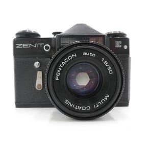 May include: A black Zenit camera with a Pentacon auto 1.8/50 lens. The camera has a textured body, a flash attachment, and various dials and buttons. The lens is marked with "Multi Coating".