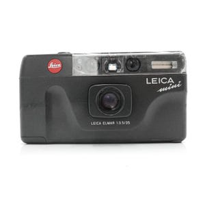 Op de afbeelding: Een zwarte Leica Mini camera met een doorzichtige flitskap. De camera heeft het Leica-logo in rood en de tekst "LEICA mini" in wit. De lens is gemarkeerd met "LEICA ELMAR 1:3.5/35".