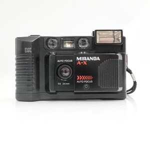 May include: A black Miranda A-X 35mm film camera with a built-in flash. The camera features the text "MIRANDA A-X" in red, along with "AUTO FOCUS" and "f/4 34mm". A wrist strap is attached.