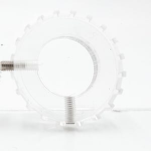 May include: A transparent, circular plastic component with a toothed outer edge and a central aperture. Two metal screws are inserted through the plastic. The object is set against a white background.