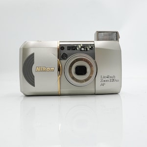 Pode incluir: Câmera Nikon Lite Touch Zoom 120 ED AF prateada. A câmera possui flash embutido e lente zoom de 38-120 mm. O logotipo da Nikon é visível na frente.