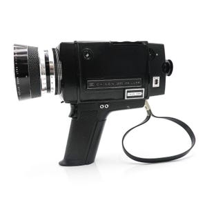 Puede incluir: Una cámara de cine Super 8 negra Chinon 1071 De Luxe con un objetivo zoom y un micrófono incorporado. La cámara tiene una correa negra y un anillo de zoom plateado.