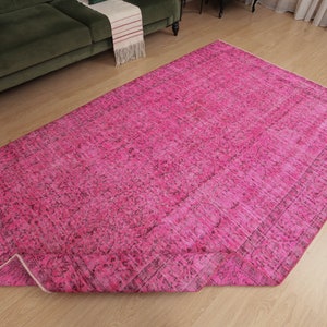 May include: A large, rectangular, hot pink rug with a distressed, vintage-inspired design. The rug features a faded, mottled pattern with darker accents, and is partially rolled up, revealing its underside. The rug is placed on a light-coloured wooden floor.