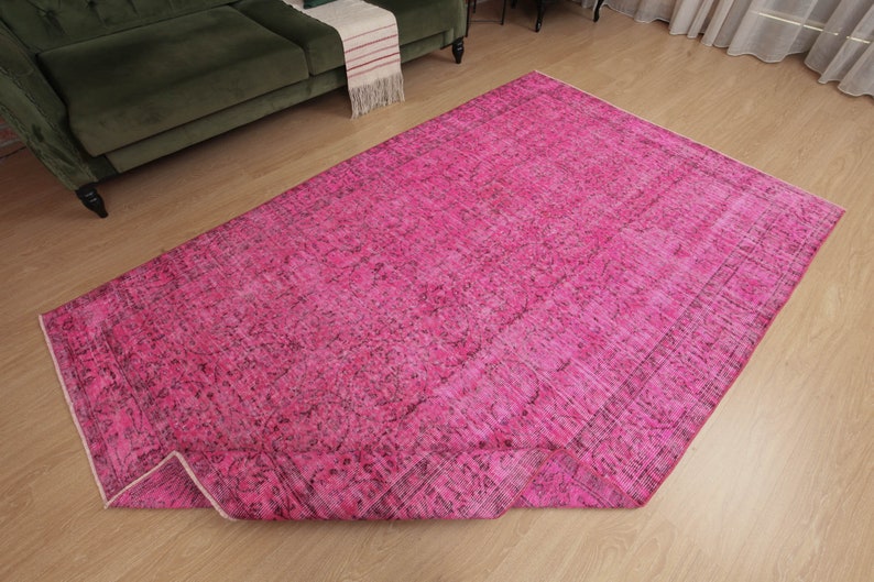May include: A large, rectangular, hot pink rug with a distressed, vintage-inspired design. The rug features a faded, mottled pattern with darker accents, and is partially rolled up, revealing its underside. The rug is placed on a light-coloured wooden floor.