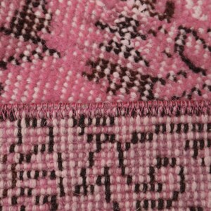 May include: A close-up view of a woven textile featuring a vibrant pink and white base with dark brown floral-inspired patterns. The fabric displays a distinct horizontal seam where two sections of the material meet, showing a stitched edge.