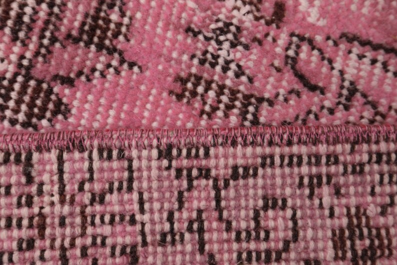 May include: A close-up view of a woven textile featuring a vibrant pink and white base with dark brown floral-inspired patterns. The fabric displays a distinct horizontal seam where two sections of the material meet, showing a stitched edge.