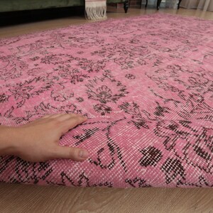 May include: A large rectangular area rug featuring a vibrant pink base color with an intricate, repeating floral pattern in dark brown. The rug is partially folded over to reveal the woven texture and the underside of the fabric construction.