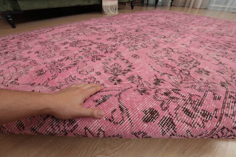 May include: A large rectangular area rug featuring a vibrant pink base color with an intricate, repeating floral pattern in dark brown. The rug is partially folded over to reveal the woven texture and the underside of the fabric construction.