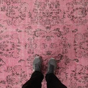 May include: A large area rug featuring a vibrant pink background with a repeating dark charcoal gray floral and damask pattern. The textile displays a distressed, weathered texture across the surface, showcasing intricate symmetrical botanical motifs.