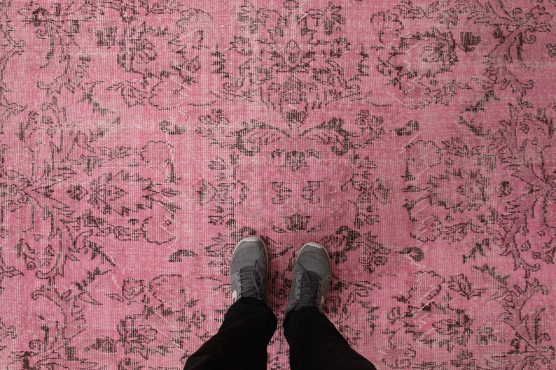 May include: A large area rug featuring a vibrant pink background with a repeating dark charcoal gray floral and damask pattern. The textile displays a distressed, weathered texture across the surface, showcasing intricate symmetrical botanical motifs.