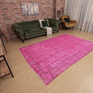 May include: A large, rectangular, hot pink rug with a distressed, vintage look dominates the room. The rug is placed on a light wood floor, with a green sofa and chair visible in the background. The room has a brick wall and a floor lamp.