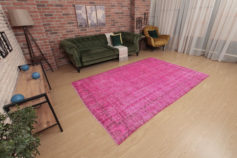 May include: A large, rectangular, hot pink rug with a distressed, vintage look dominates the room. The rug is placed on a light wood floor, with a green sofa and chair visible in the background. The room has a brick wall and a floor lamp.