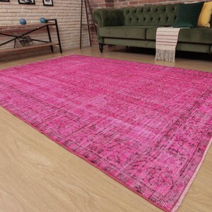 May include: A large, rectangular, hot pink rug with a distressed, vintage appearance. The rug has a faded, textured design with darker pink and black accents. It is placed on a light-coloured wooden floor.