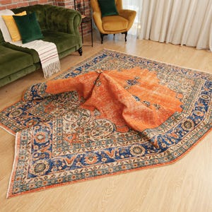 May include: A large rectangular area rug featuring a vibrant orange central field with a distressed, faded texture. The rug is bordered by intricate floral and geometric patterns in deep blue, cream, and muted green tones with fringed edges.