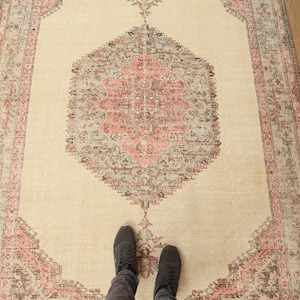 May include: A vintage rug with a cream background and a large, intricate, pink and gray floral design in the center. The rug has a faded, distressed look and a pink and gray border.