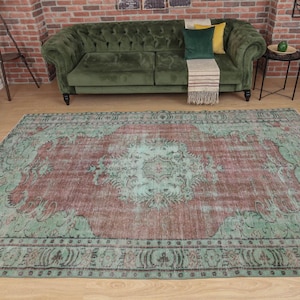 May include: Large, rectangular area rug with a distressed, vintage look. The rug features a central medallion design in shades of burgundy and teal, surrounded by a teal border. The rug is placed on a wooden floor, with a green sofa and yellow armchair in the background.