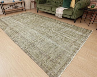 May include: A large rectangular area rug featuring a faded olive green and beige color palette with an intricate, repeating floral and vine pattern throughout the center and a wide, detailed border framing the entire piece on a light wood floor.