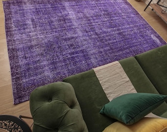 May include: A purple vintage rug with a distressed look, placed on a wooden floor. A green sofa with a white throw pillow and a green velvet pillow is partially visible in the foreground.