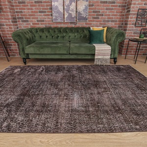 Op de afbeelding: Een donkerbruin, verweerd vloerkleed met een subtiel patroon. Het kleed bevindt zich in een woonkamer met een groene bank en een bakstenen muur op de achtergrond.