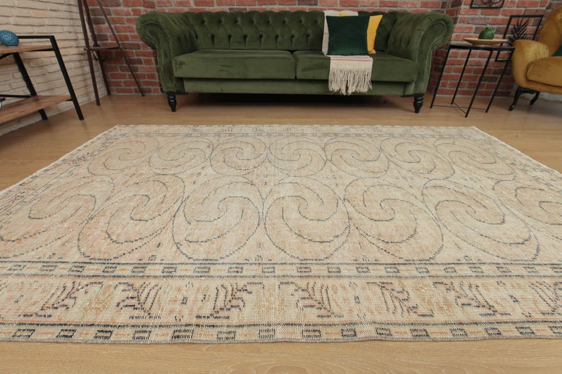 May include: A beige area rug with a swirling pattern in a darker beige color. The rug has a border with a repeating geometric design.