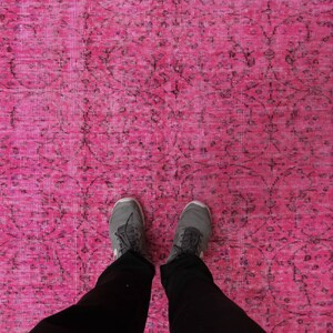 May include: A bright pink rug with a detailed, dark pattern. The rug's design includes floral and geometric shapes. The image shows a person's feet and legs, wearing grey trainers and black trousers, standing on the rug.