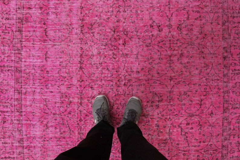 May include: A bright pink rug with a detailed, dark pattern. The rug's design includes floral and geometric shapes. The image shows a person's feet and legs, wearing grey trainers and black trousers, standing on the rug.