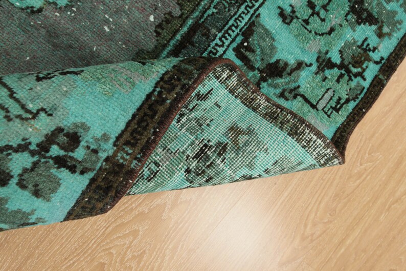 May include: Close-up of the edge of a teal and black patterned rug with a brown fringe. The rug is folded over and the underside is visible.