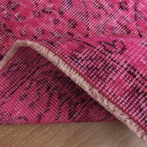 May include: Close-up of a vibrant pink rug with a detailed, patterned design. The rug is folded over, revealing its textured surface and fringed edge. The colour palette includes shades of pink and dark burgundy.