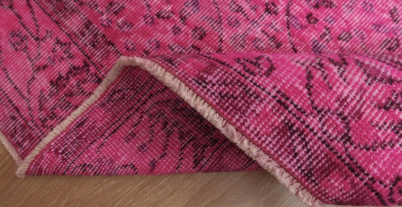 May include: Close-up of a vibrant pink rug with a detailed, patterned design. The rug is folded over, revealing its textured surface and fringed edge. The colour palette includes shades of pink and dark burgundy.