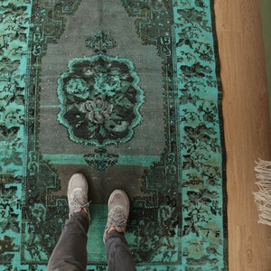 May include: A teal and black vintage rug with a floral pattern. The rug is laid out on a wooden floor with a person's feet standing on it.