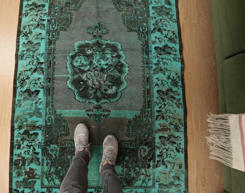 May include: A teal and black vintage rug with a floral pattern. The rug is laid out on a wooden floor with a person's feet standing on it.