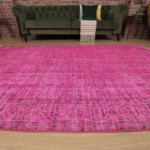 May include: A large, rectangular, cerise rug with a distressed, vintage-inspired design. The rug has a faded, textured appearance with darker accents along the edges and within the pattern. The rug is placed on a light wood floor.