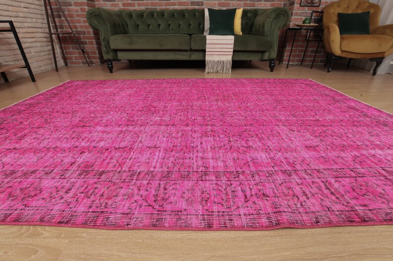May include: A large, rectangular, cerise rug with a distressed, vintage-inspired design. The rug has a faded, textured appearance with darker accents along the edges and within the pattern. The rug is placed on a light wood floor.