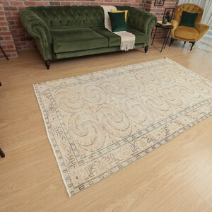 May include: A cream-colored rug with a geometric pattern of swirls and lines. The rug has a light brown border and is laid out on a wooden floor.
