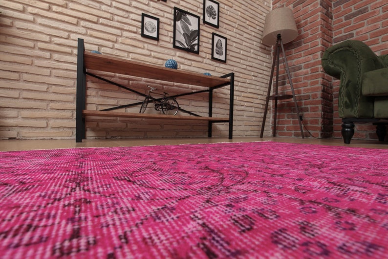 May include: A bright pink rug with an intricate design fills the lower portion of the image. The room features a brick wall, wooden shelving, and a floor lamp. The overall aesthetic suggests a warm and inviting space.