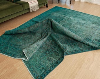 Puede incluir: Una alfombra rectangular grande de color verde azulado profundo con un patrón tradicional desgastado y descolorido que presenta intrincados motivos florales y geométricos. La alfombra está parcialmente doblada, revelando su textura y el reverso.