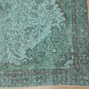 May include: A distressed, rectangular rug with a faded, floral pattern in shades of teal, green, and charcoal. The rug has a border with a darker, more defined design. The rug's design is reminiscent of vintage or antique styles.