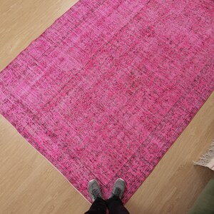May include: A rectangular, hot pink rug with a distressed, vintage appearance. The rug has a darker border and is placed on a light wood floor. The rug's dimensions are not visible.